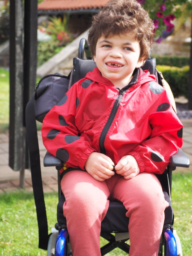 Pudding smiling widely seated in his wheelchair wearing a bright red coat with ladybird spots.