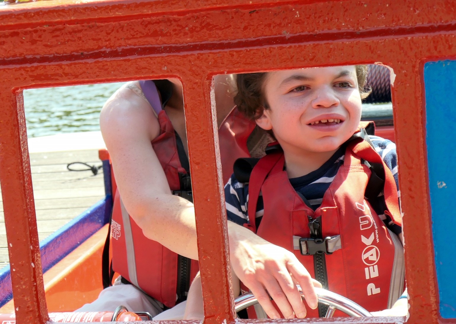 Pudding looking through the front window of a boat holding onto the steering wheel. I am sitting by him, arm just visible guiding the wheel too.