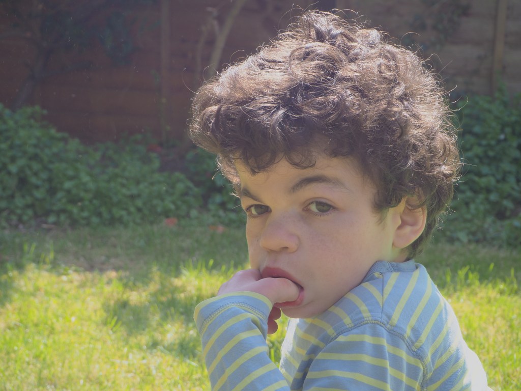 A close up photo of boy with curly hair with his thumb in his mouth. He is not smiling.