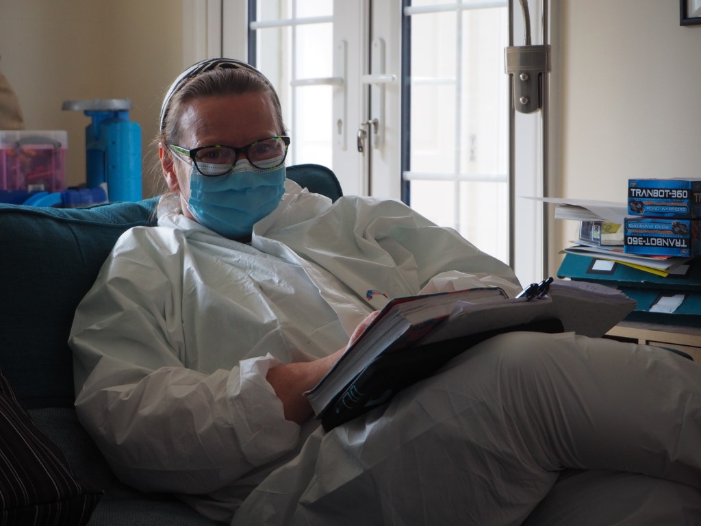 Nurse sitting on sofa with notebooks, wearing mask and white coverall PPE. She has smiling eyes.